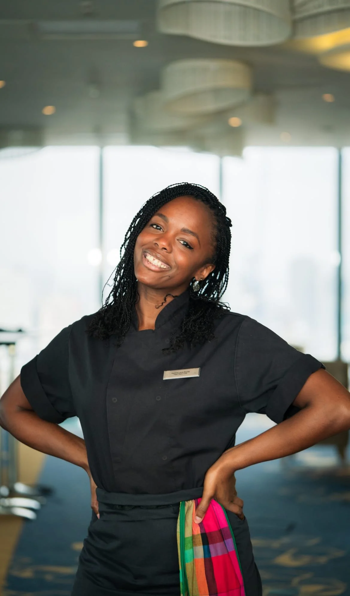 Smiling woman in a black chef uniform with a colorful towel tucked into her waistband, standing confidently with hands on hips in a modern, well-lit restaurant or hotel lobby.