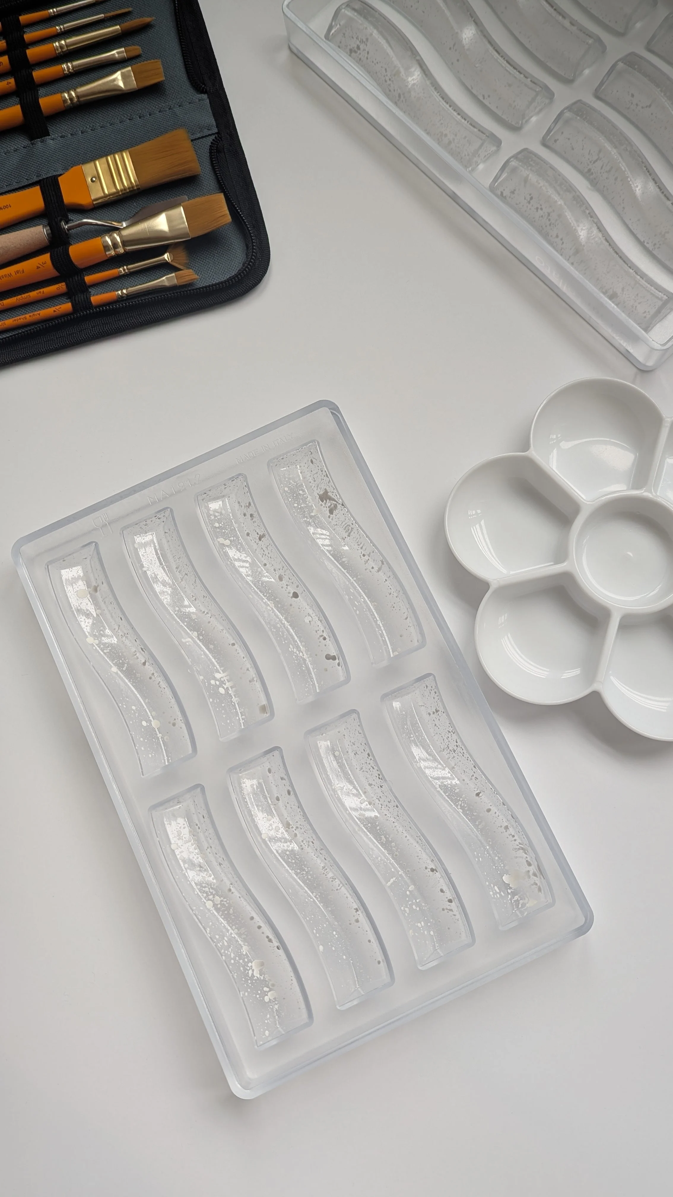 Empty polycarbonate mould and palette tray used for painting or crafting, with a white ceramic paint palette with multiple wells, and a set of paintbrushes in a black case.