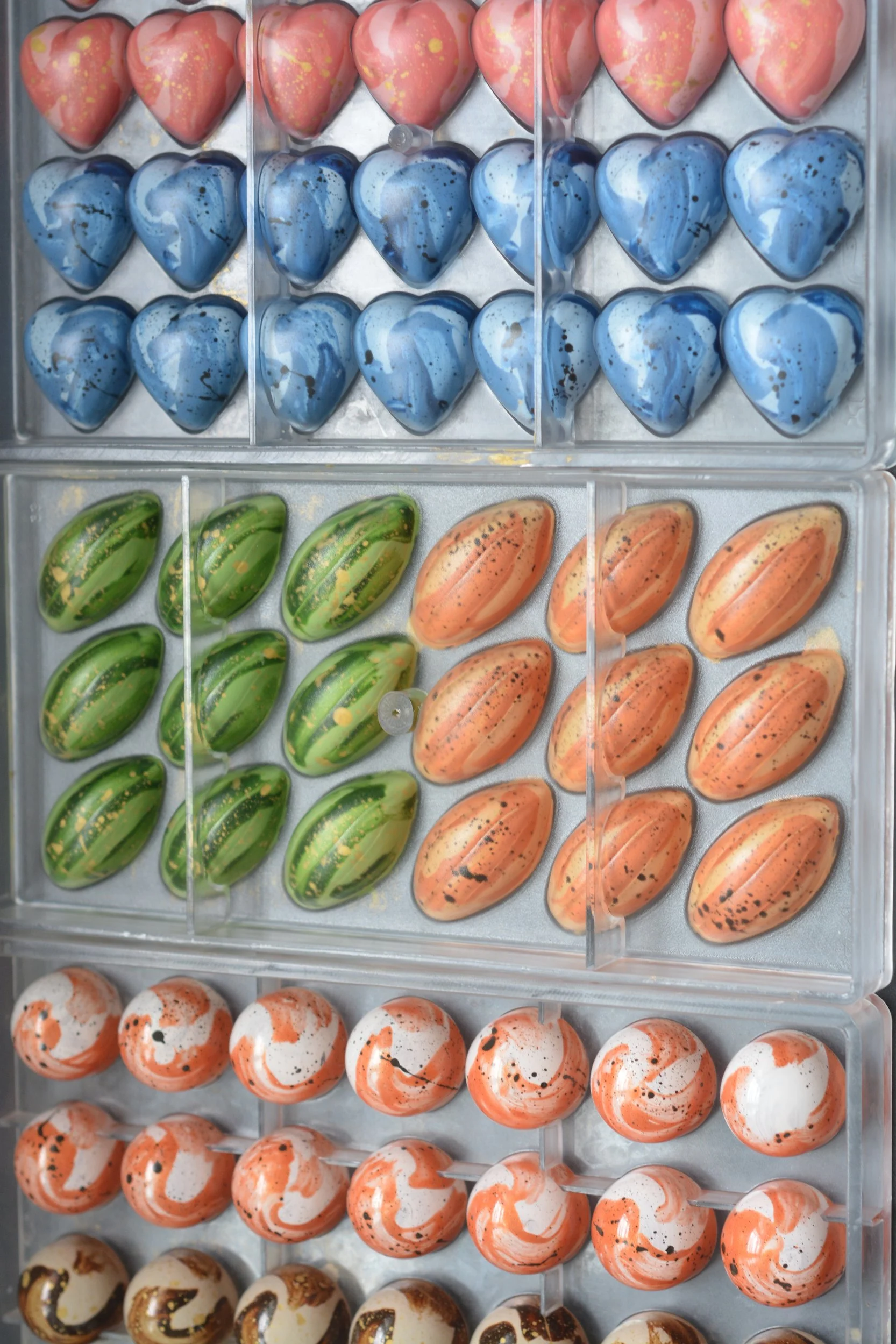 A display case filled with colorful, decorative chocolate candies shaped like hearts, eggs, and spheres, decorated with marbled and speckled patterns in pink, blue, green, orange, white, and gold.