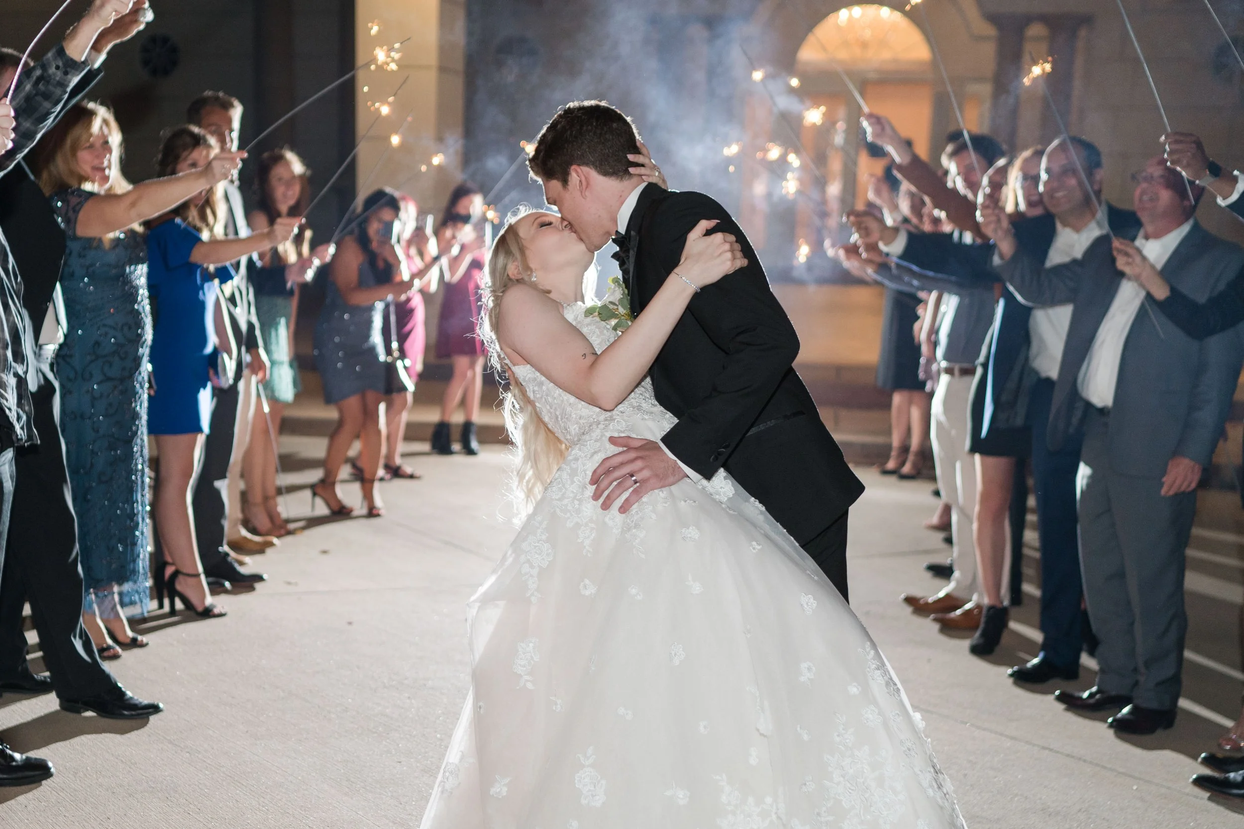 A newlywed couple sharing a kiss during their wedding celebration while friends and family hold sparklers surrounding them.