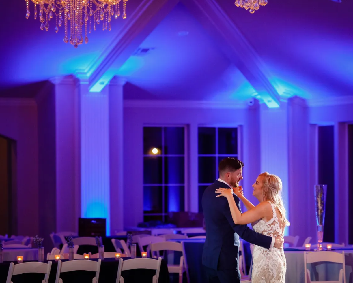 Elegant banquet room with round tables covered in blue tablecloths, decorated with tall floral centerpieces of pink and white roses adorned with hanging crystals. Crystal chandelier and ornate moldings on the ceiling and walls add to the luxurious atmosphere.
