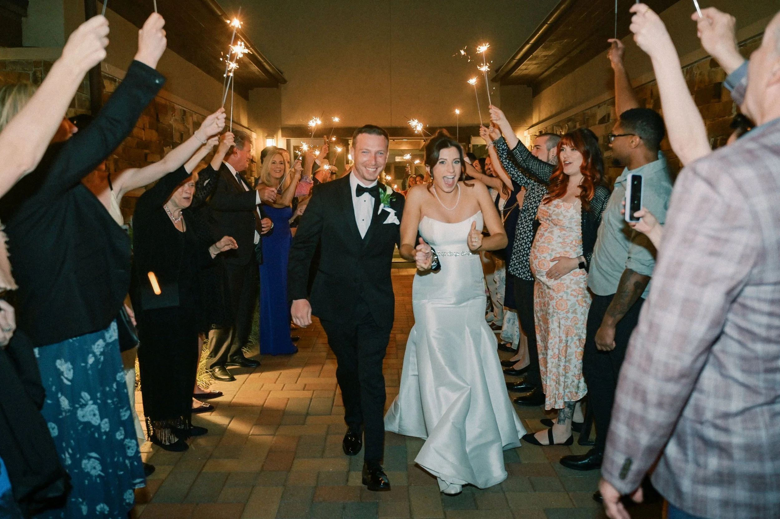 Bride and groom walking through a tunnel of guests holding sparklers at night during a wedding celebration.