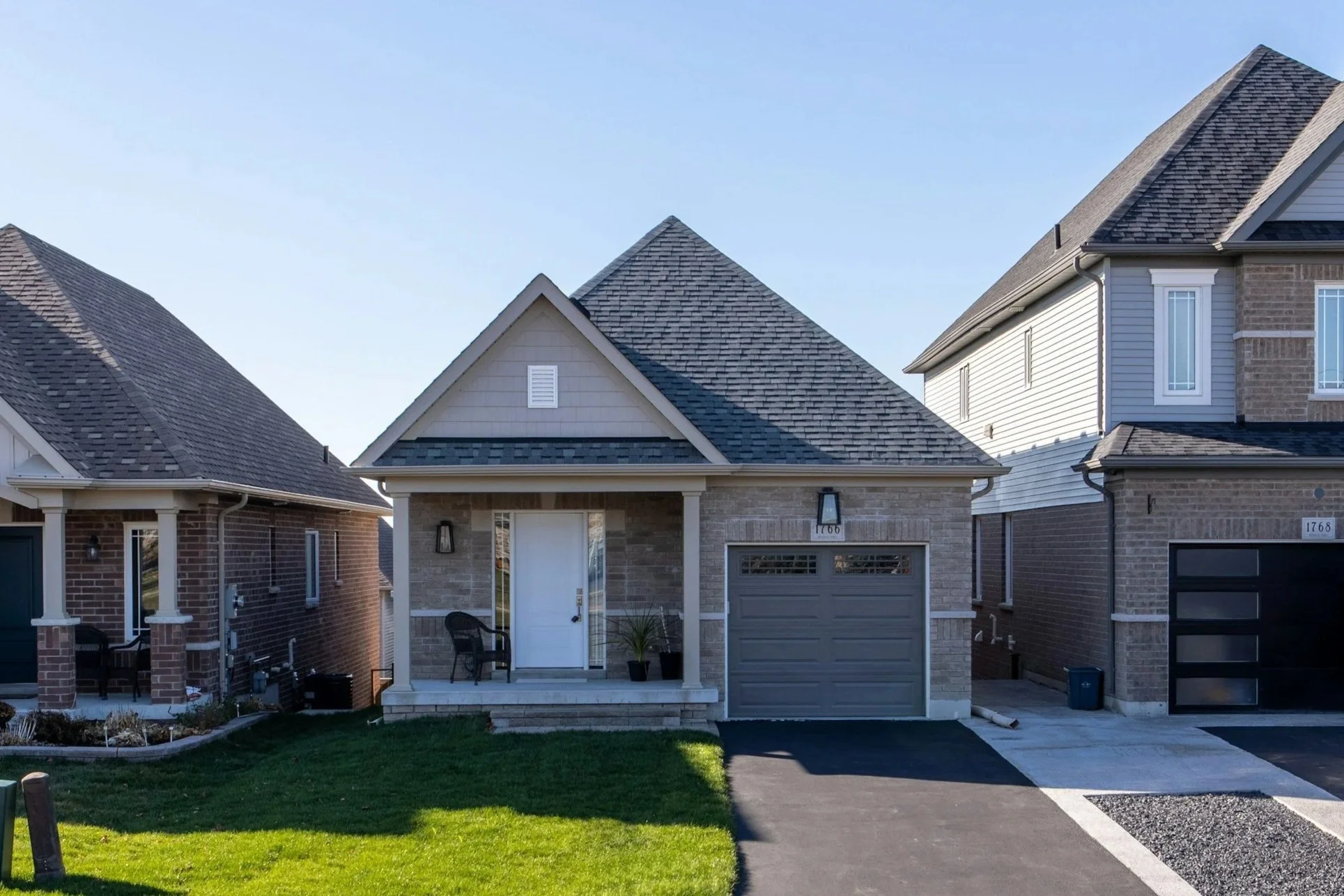 Front view of a modern house with brick and siding exterior, gray roof, front porch with chairs, and a garage door, neighboring houses on each side, a black mailbox, and a sunny day.