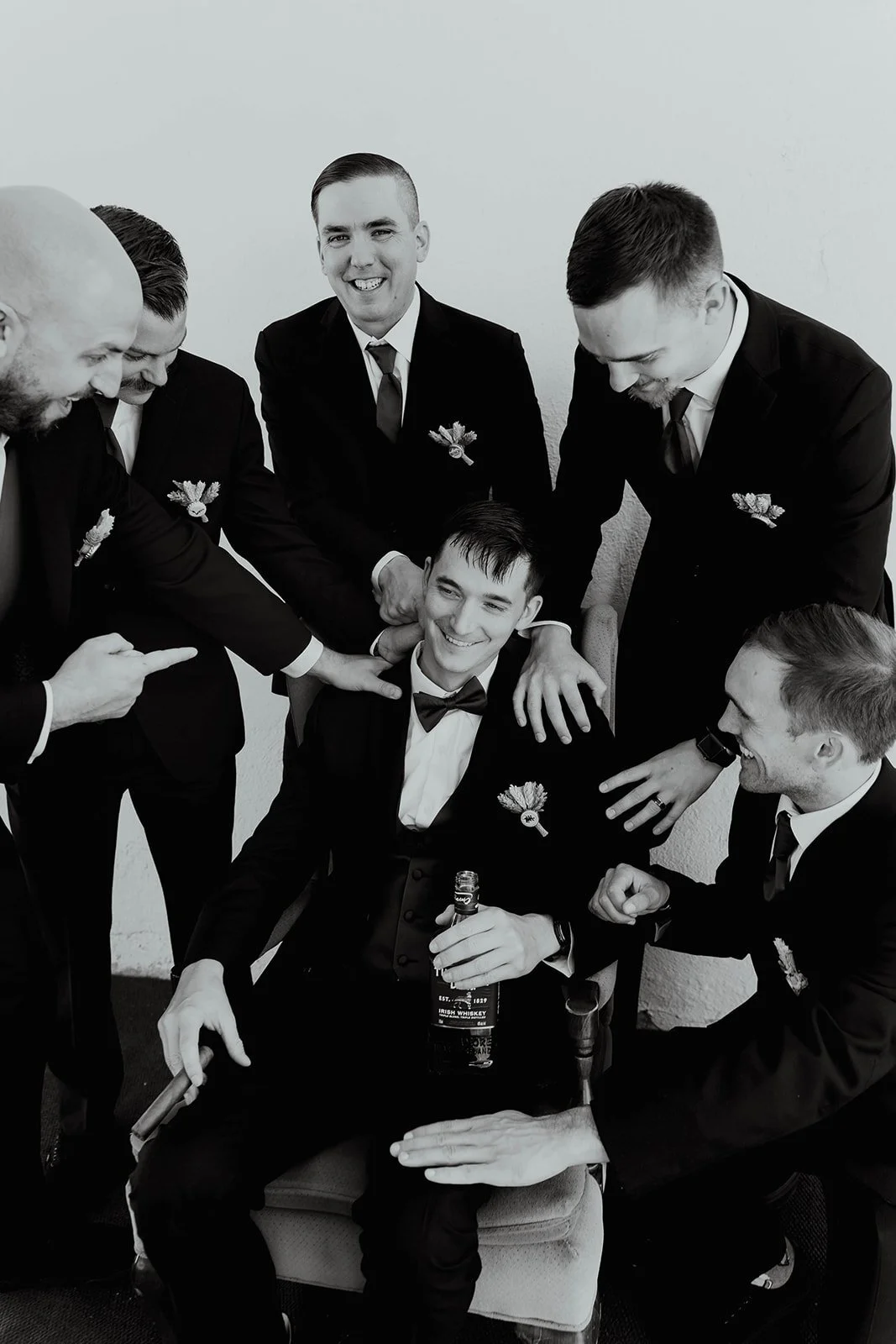 A group of men in tuxedos surrounding a seated man, who is holding a bottle, smiling, and wearing a bow tie, while the others are playfully touching his head and shoulder.