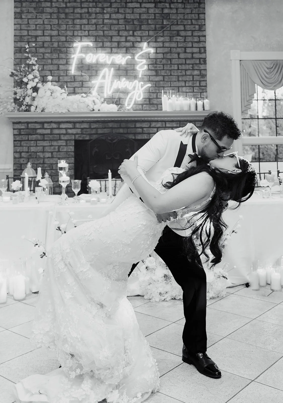 A bride and groom sharing a dance at their wedding reception, with a brick fireplace, floral decorations, candles, and a neon sign reading 'Forever & Always' in the background.