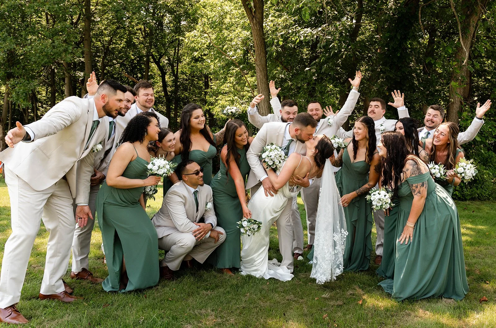 A group of wedding guests and the bride and groom are laughing and celebrating outdoors in a wooded area. The bride and groom are kissing, with the groom holding the bride. The guests are wearing green dresses and beige suits, and some hold white bou