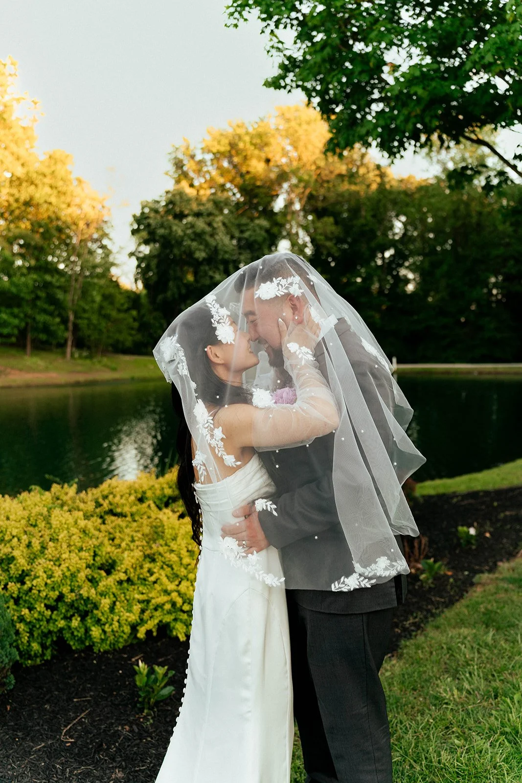 A bride and groom sharing a close moment under a veil outdoors near a lake with trees in the background.