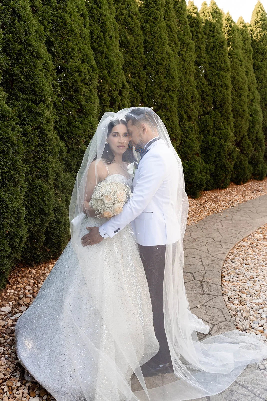 A bride and groom standing close together under a white veil, with green shrubs in the background. The bride is holding a bouquet and wearing a white wedding dress, and the groom is in a white tuxedo jacket with black pants.
