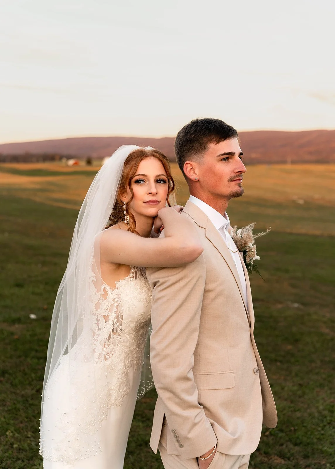 A bride and groom posing outdoors during sunset, with the bride resting her arm on the groom's shoulder. The bride wears a lace wedding dress with a veil, and the groom wears a beige suit with a white shirt. The landscape features an open field with