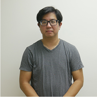 A man with black hair and glasses, wearing a gray T-shirt, standing against a plain off-white wall.