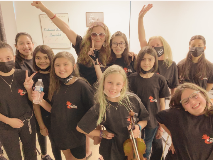 Group of young girls and a woman, some holding a water bottle and a violin, posing happily together indoors.