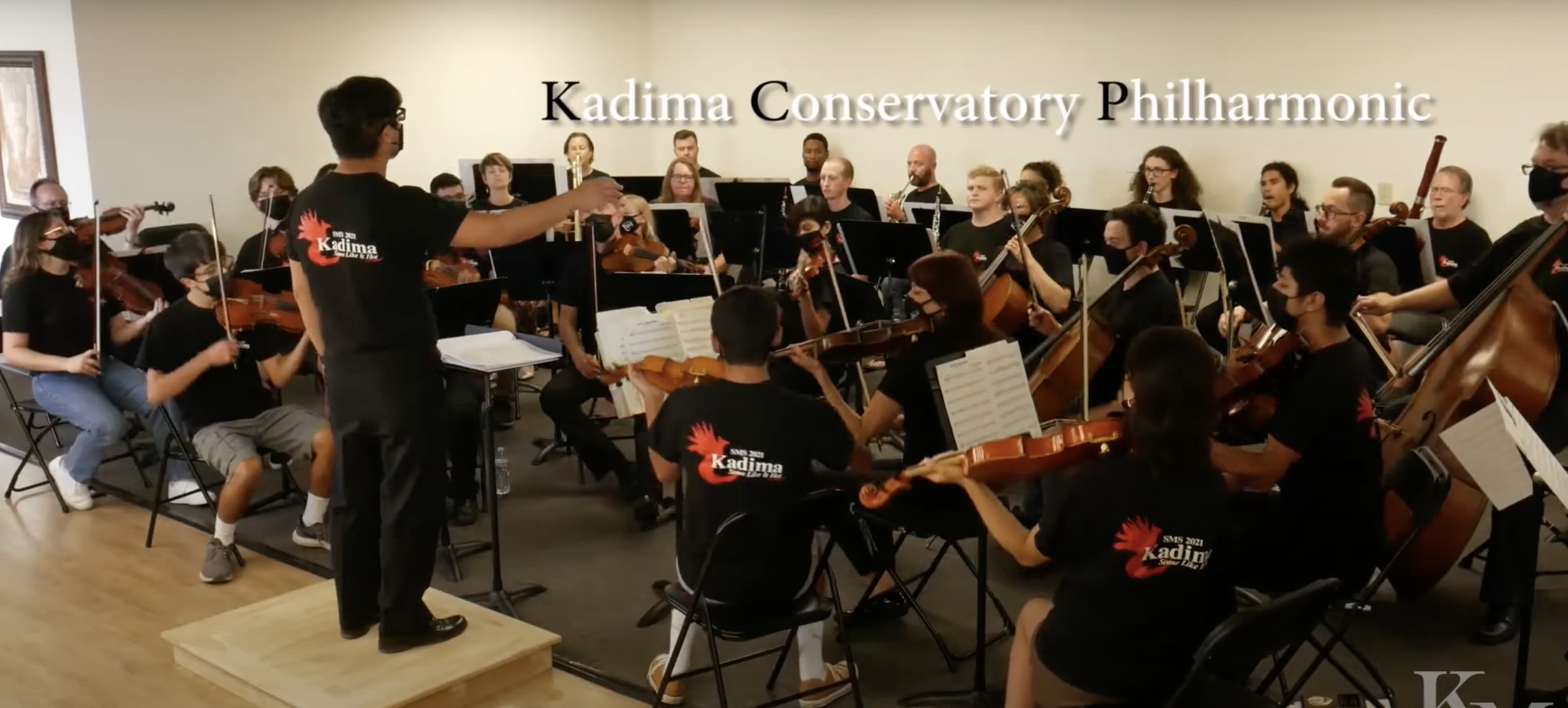 Orchestra rehearsing in a concert hall at Kadima Conservatory Philharmonic, with a conductor standing on a small platform. The musicians are playing various string instruments, and some are wearing black shirts with the Kadima logo.