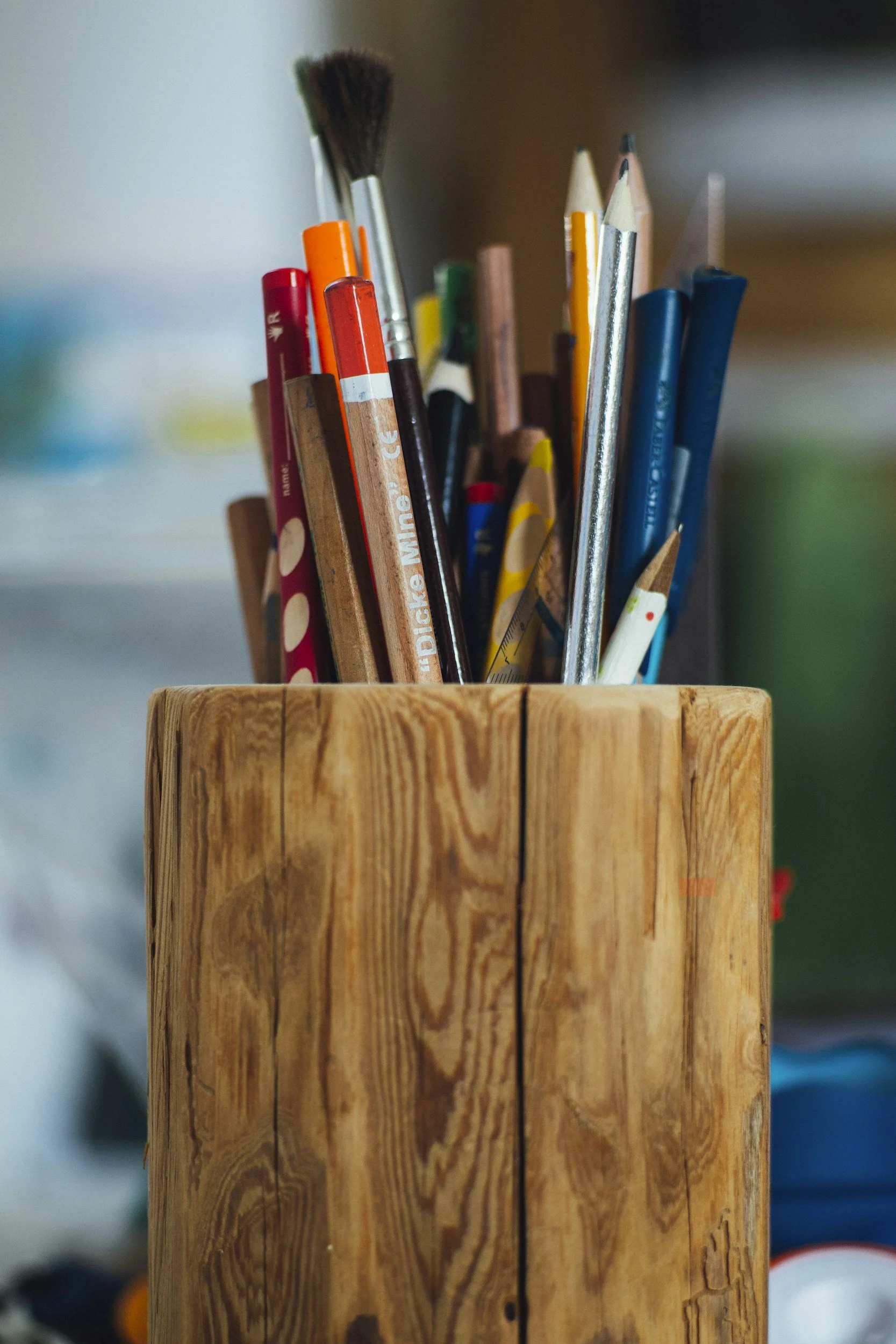 A wooden container holding various colored pencils, paintbrushes, and drawing tools.