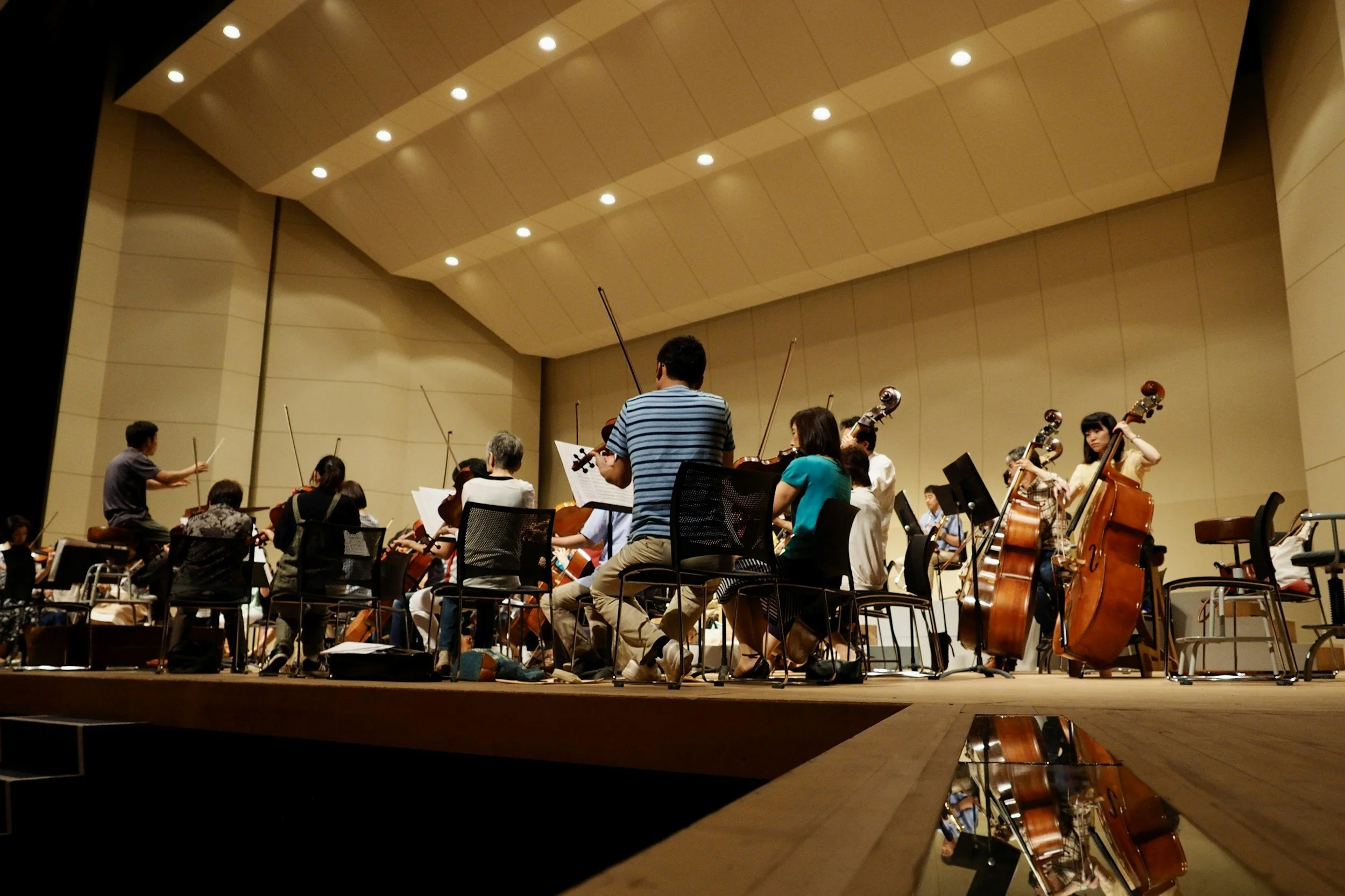 Orchestra members practicing on stage with strings, using music stands, under warm lighting in a concert hall