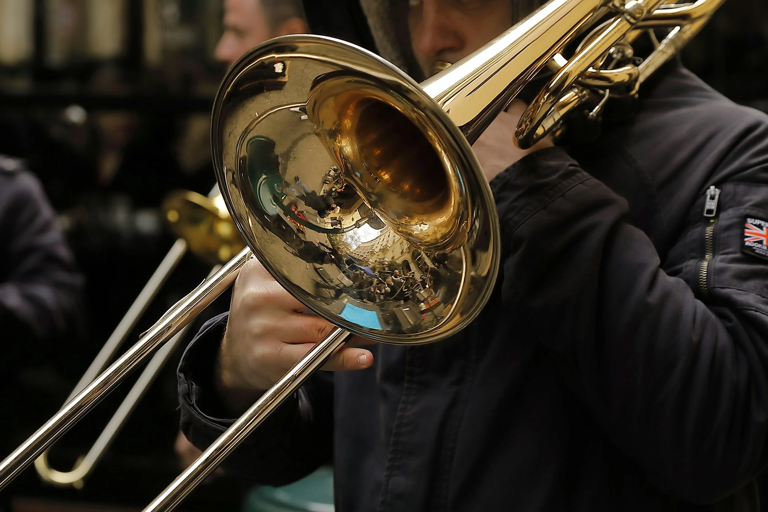 A person playing a brass trombone, with the reflection of a street scene visible on the instrument's bell.
