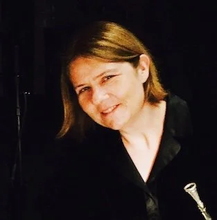 A woman with chin-length brown hair smiling, wearing a black shirt against a black background
