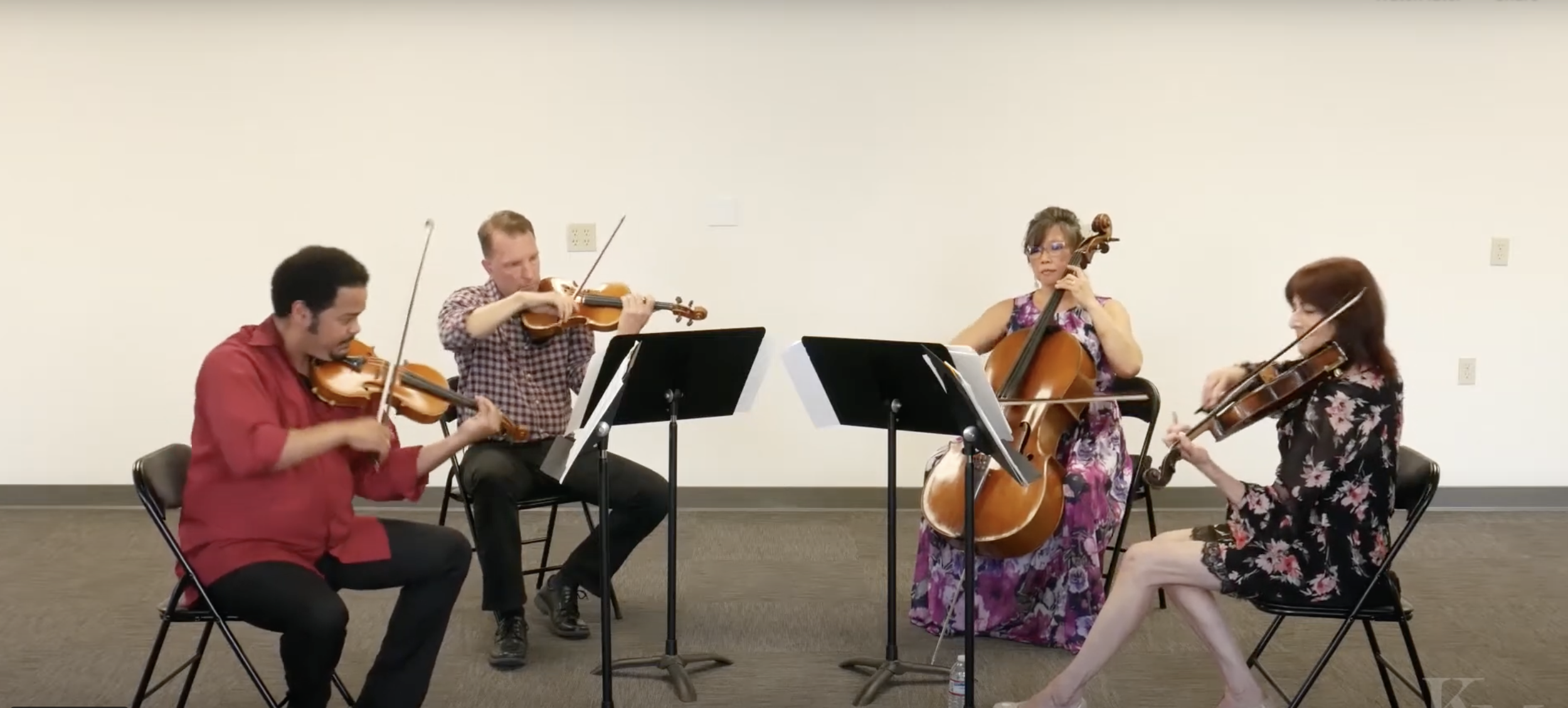Four musicians playing strings instruments: two women and two men, sitting in a room with a plain white wall, music stands, and chairs, performing together.