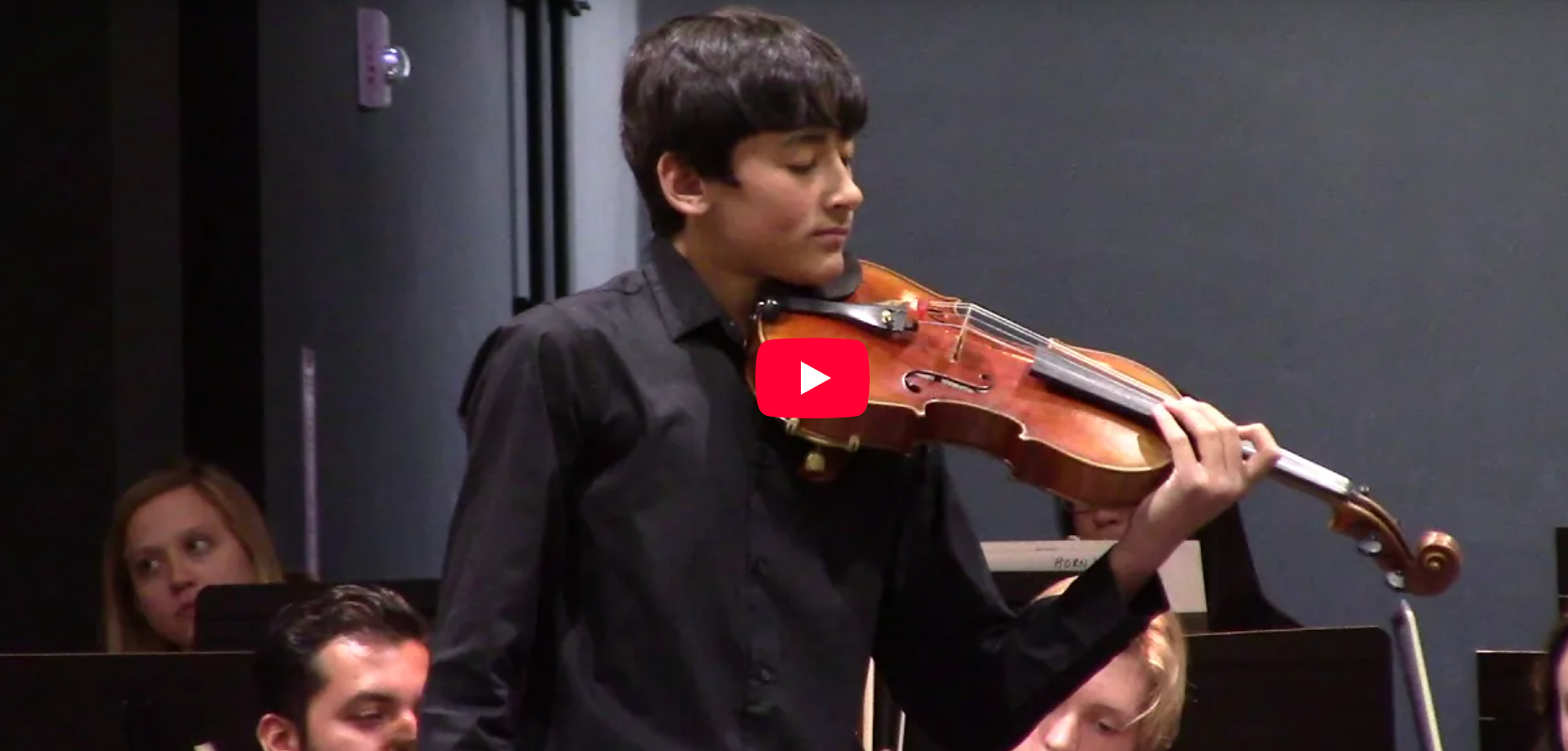 A young man playing the violin in a concert hall, with other musicians visible in the background.