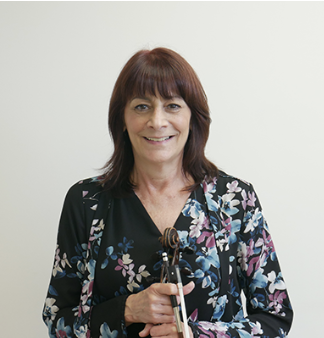 A woman with shoulder-length brown hair holding a violin and bow, smiling, wearing a floral-patterned black shirt, standing against a plain white wall.
