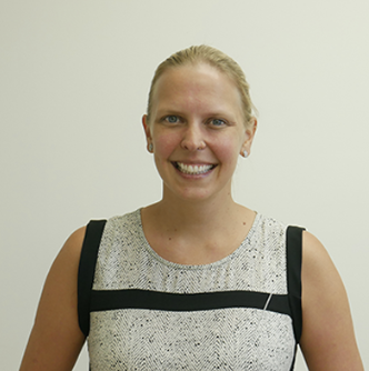 A smiling woman with blonde hair in a ponytail, wearing a sleeveless light-colored top with black accents, standing against a plain background.
