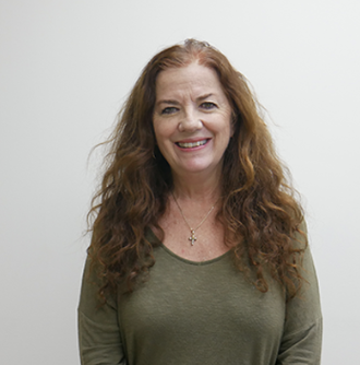 A woman with long curly red hair, wearing a green top and a necklace, smiling at the camera against a white background.