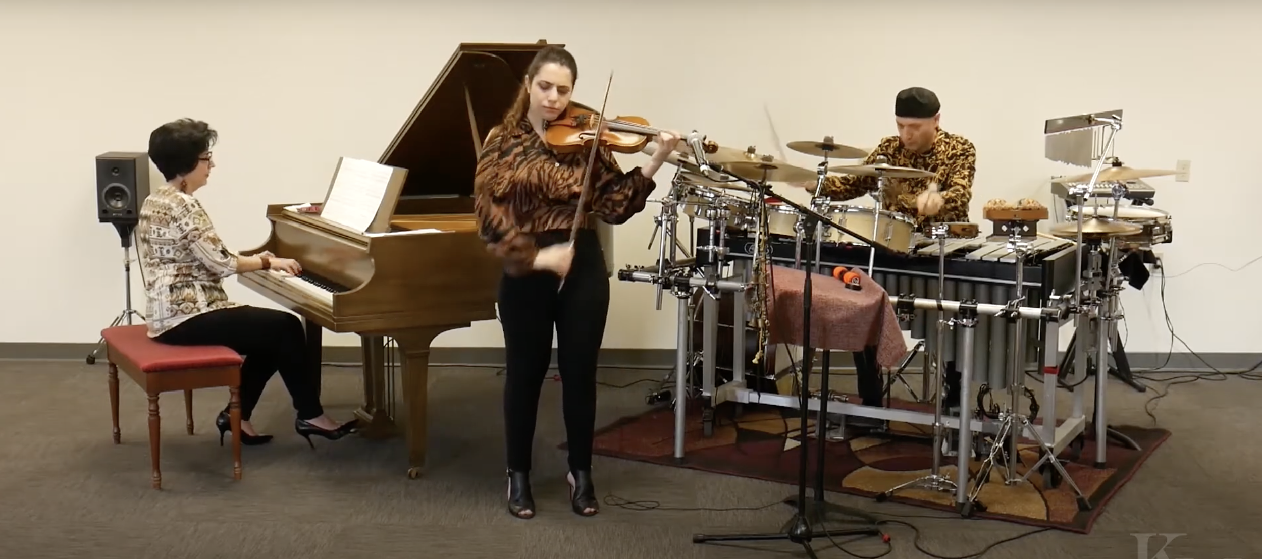 Three musicians performing in a room: a woman playing the piano on the left, a woman playing the violin in the center, and a man playing percussion instruments on the right.