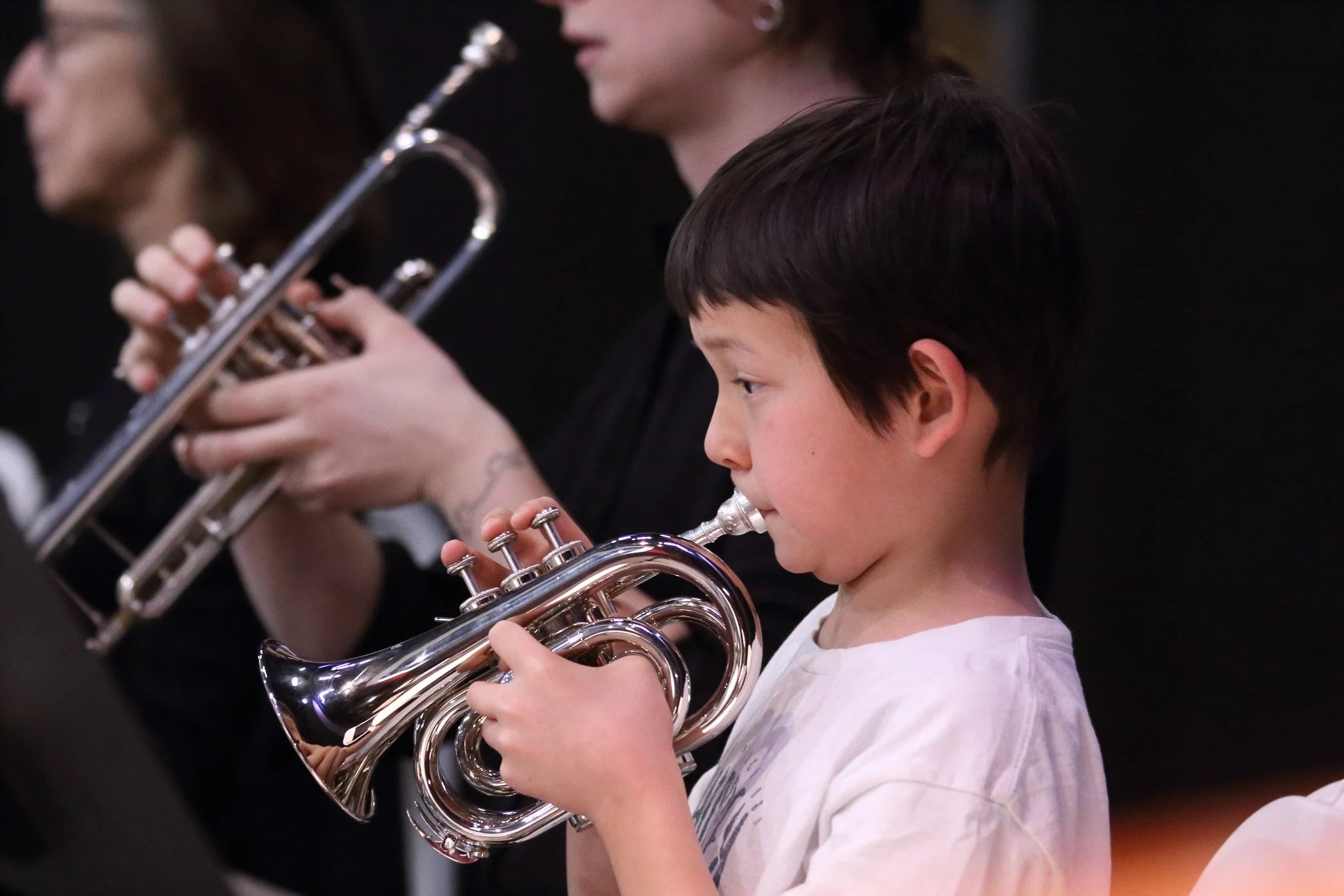 A young boy playing a silver trumpet during a music class or rehearsal, with adult musicians also playing instruments in the background.