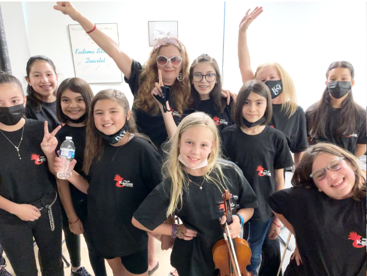 Group of young girls and a woman in black T-shirts, some wearing face masks and glasses, posing together indoors in front of a whiteboard. One girl is holding a violin. They are smiling and making peace signs or raising their hands.