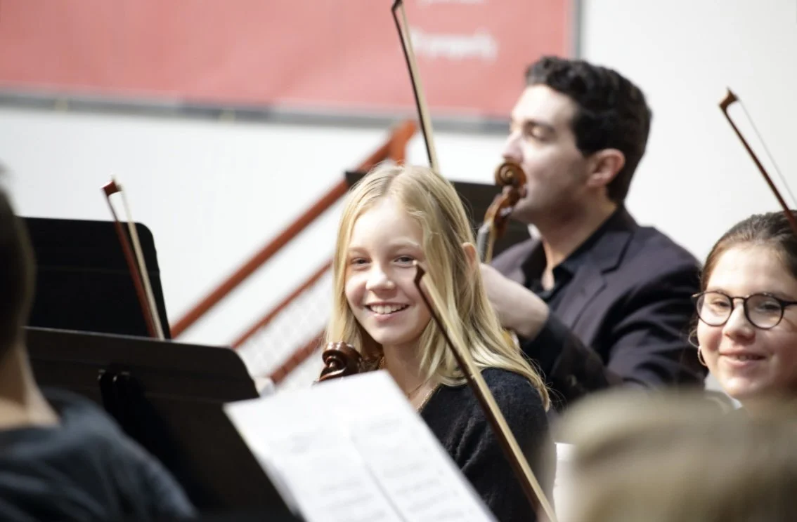 Young girl smiling during an orchestra rehearsal, surrounded by musicians, with music stands and instruments visible.