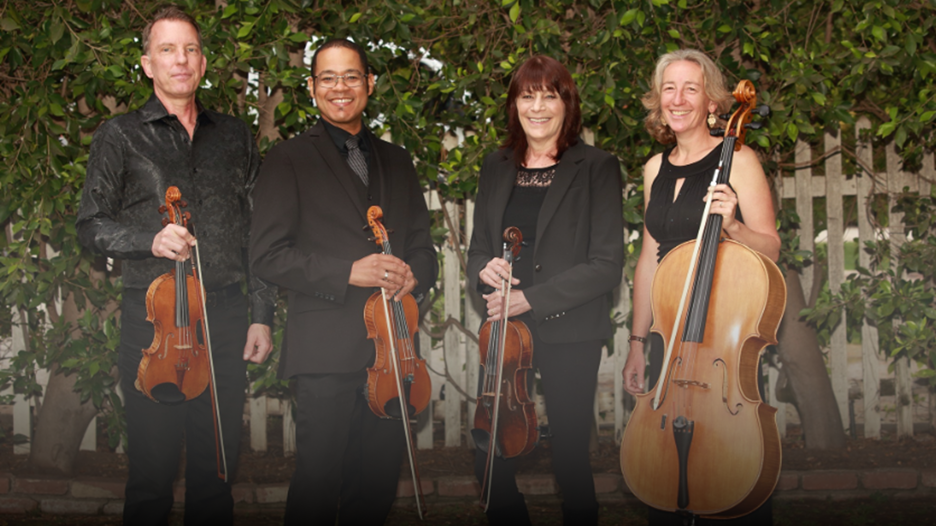 Four musicians holding string instruments, including violins, a viola, and a cello, standing outdoors in front of a background of trees and a white picket fence.