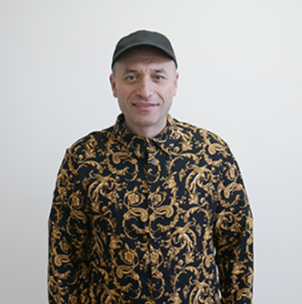 A man wearing a black cap and a black and gold patterned shirt, standing against a plain white background.
