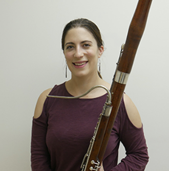 Woman holding bassoon, smiling, against a plain background.