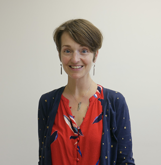 A woman with short brown hair smiling, wearing a red blouse and a navy cardigan with tiny gold dots, standing against a plain white wall.