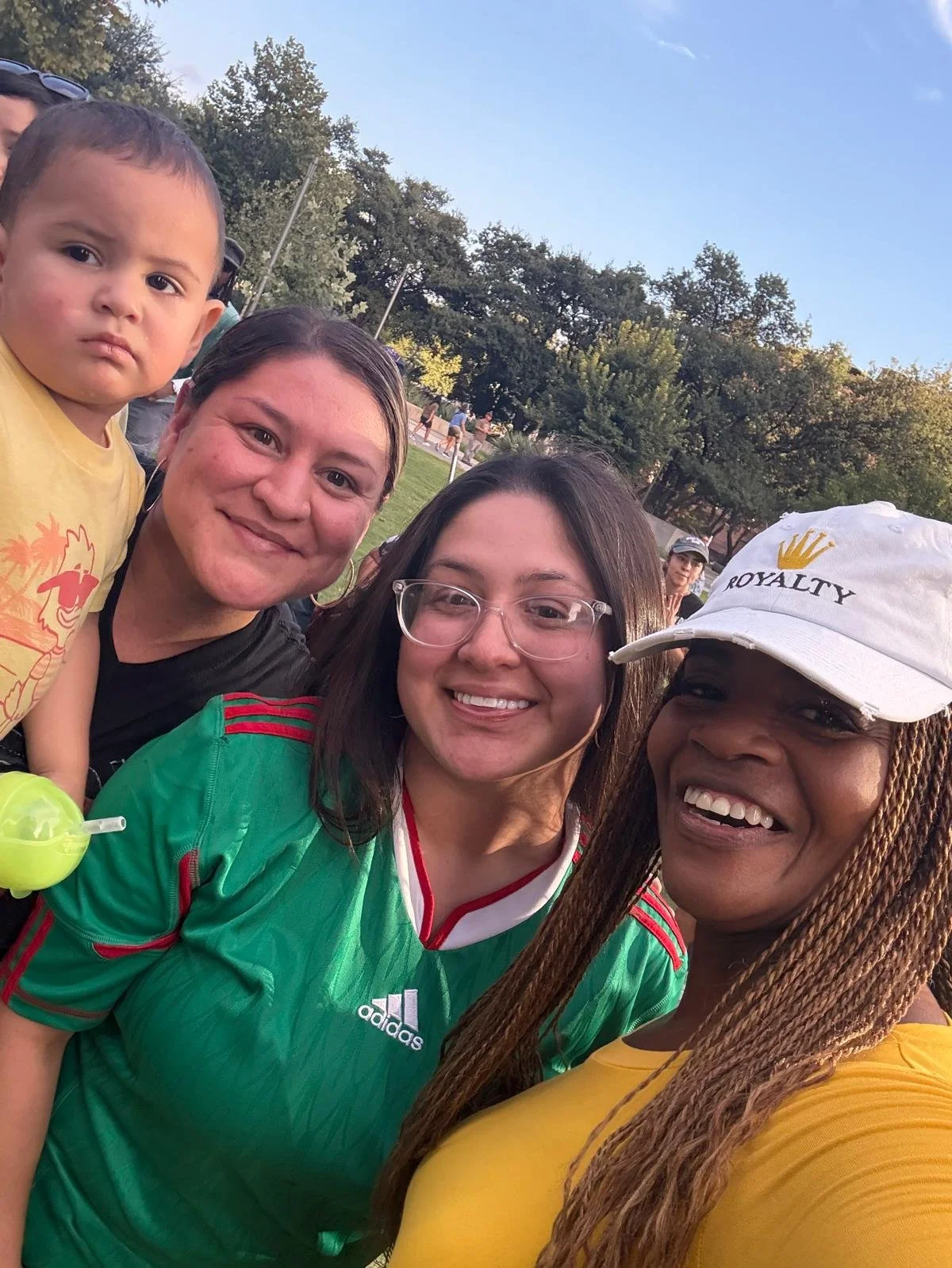 Group of four women and a young boy outdoors in a park, smiling and taking a selfie. Trees and a blue sky are in the background.