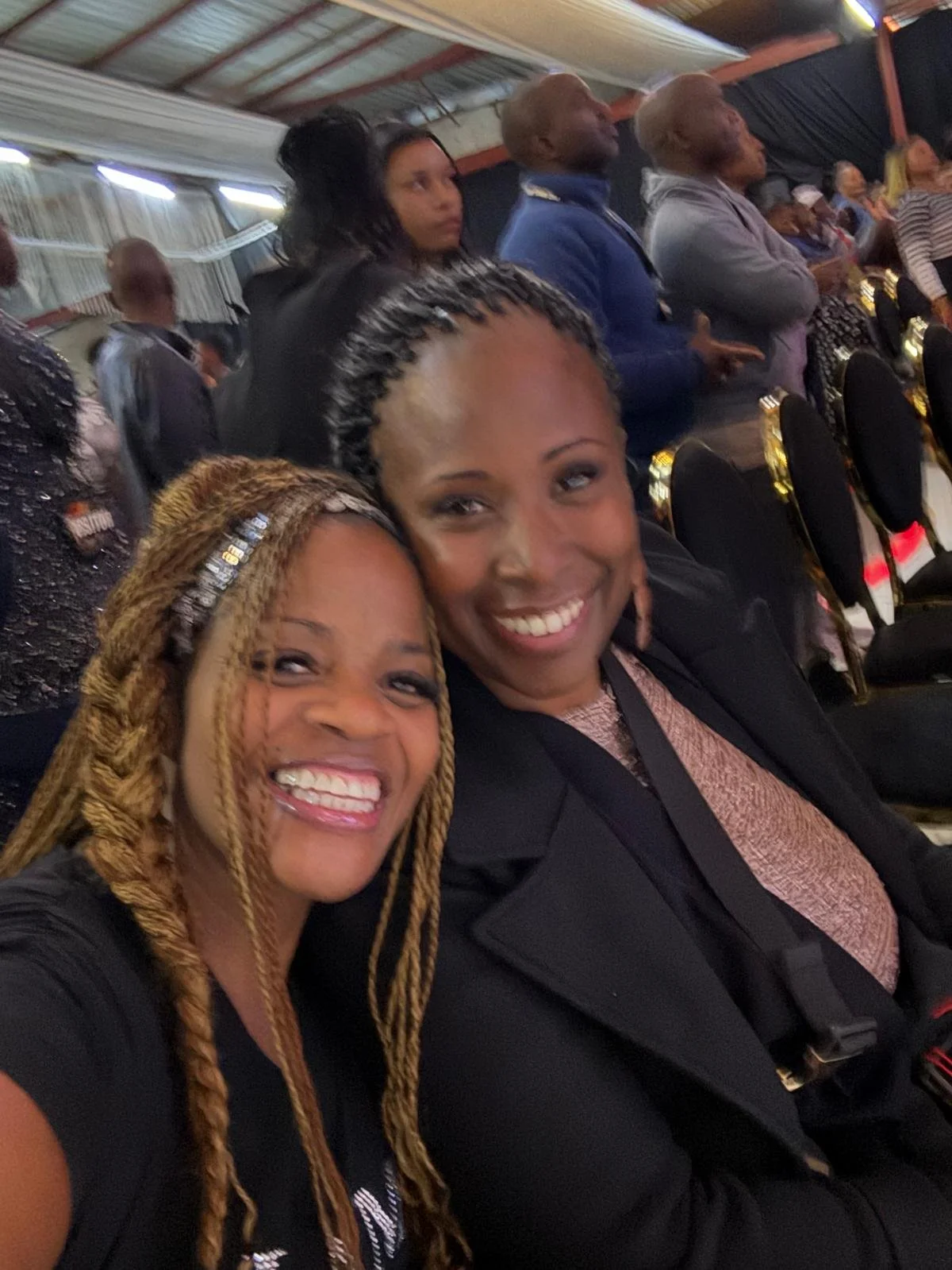 Two women smiling for a selfie at an indoor event with many people seated behind them.