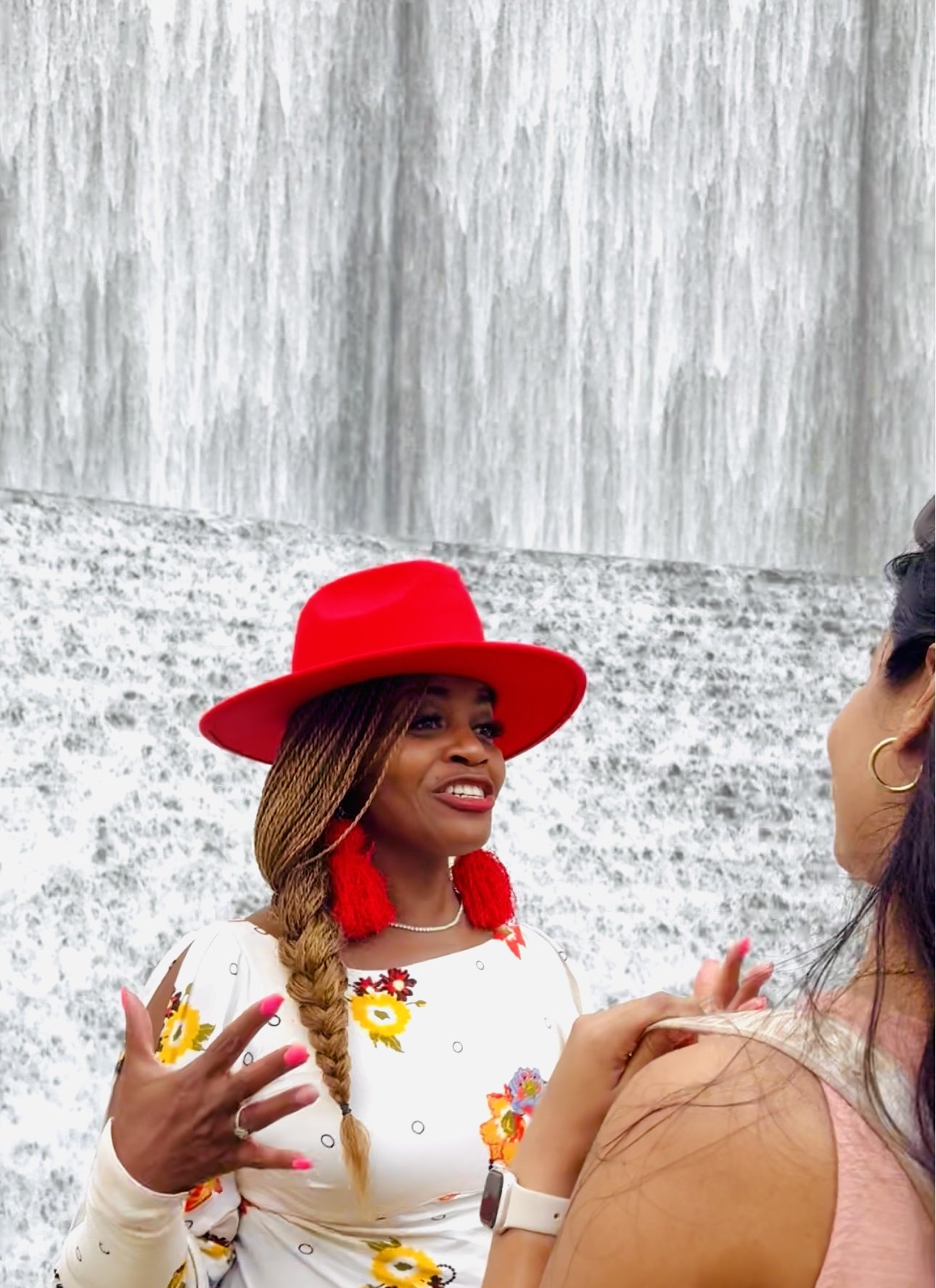 Woman in a floral dress and a large red hat with red earrings, talking to another woman near a waterfall.