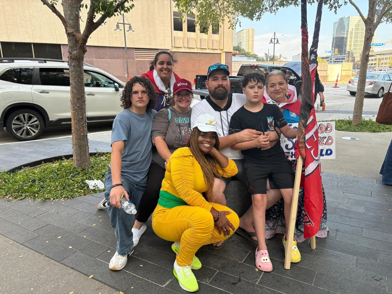 Group of people posing outdoors near a tree and parked cars, some holding signs and flags, on a city sidewalk.