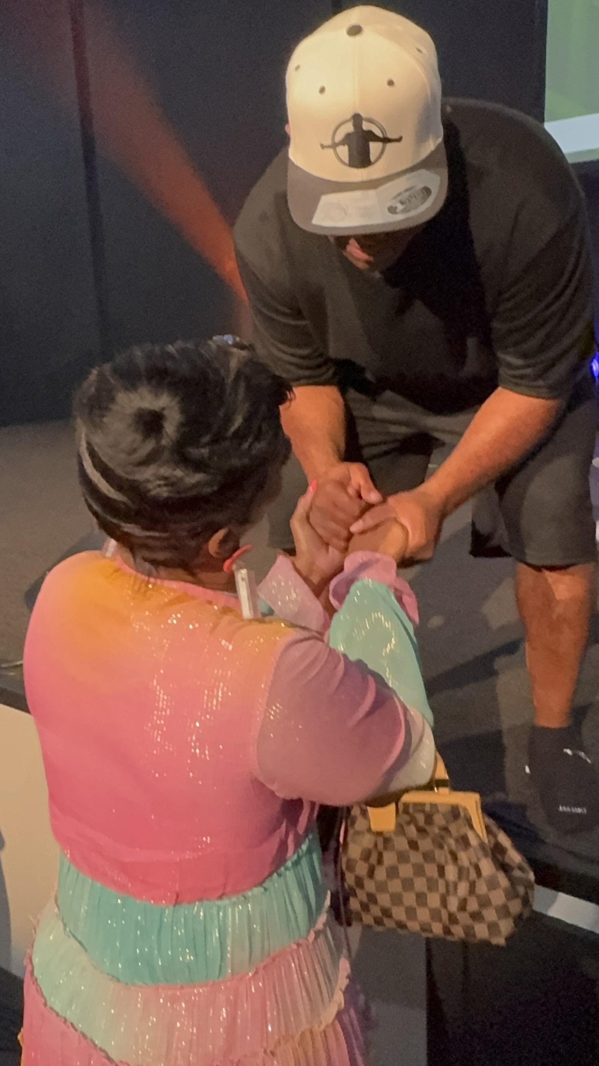A person in colorful clothing and a cap is arm wrestling with another person wearing a black shirt, shorts, and a cap, in a dimly lit indoor setting.
