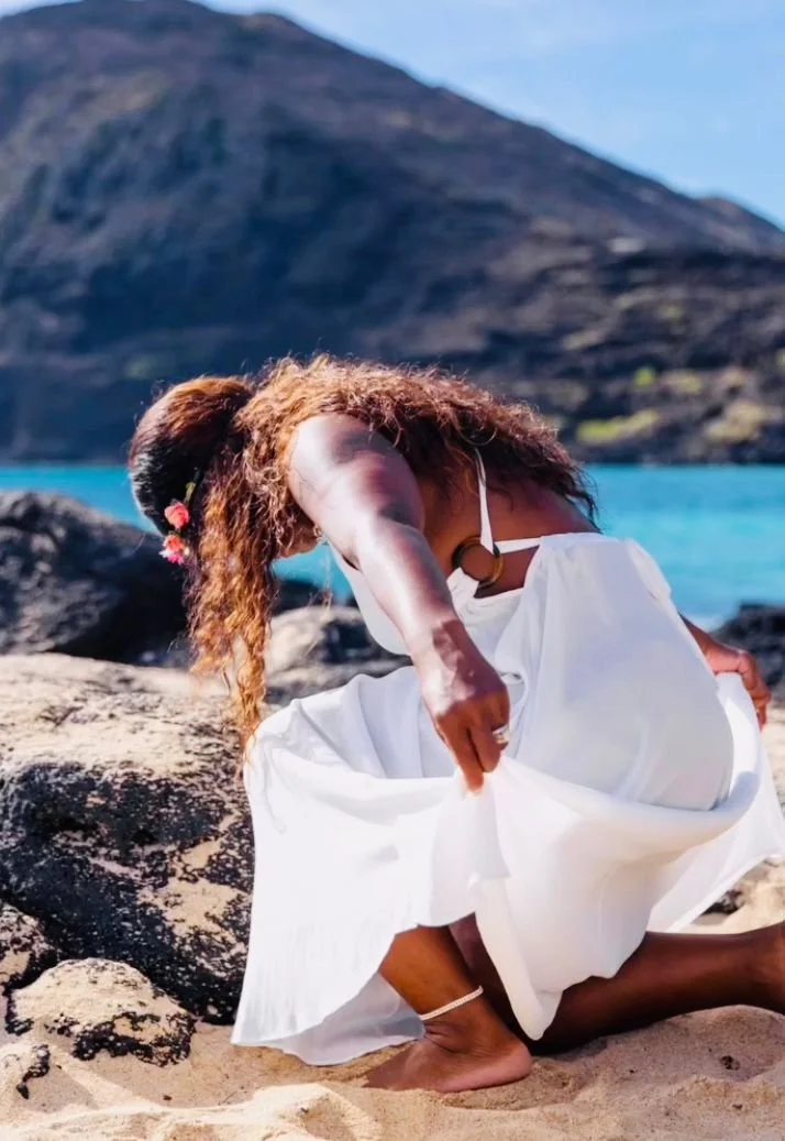 A woman in a white dress and face mask crouches on a beach with rocks, with a mountain and blue water in the background.