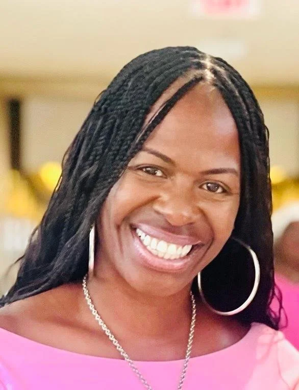 A woman with long black braided hair smiling, wearing a pink top, hoop earrings, and a silver necklace.