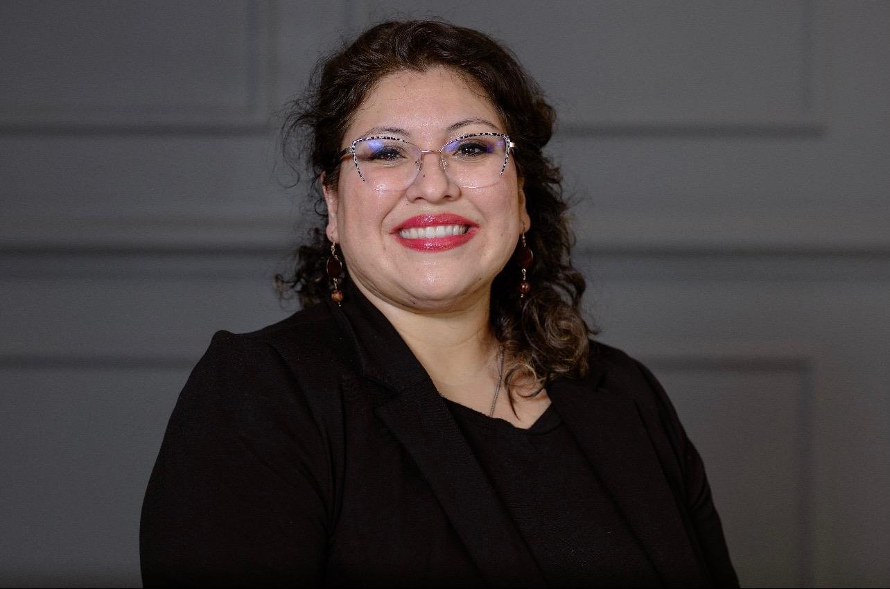 A woman with dark curly hair, glasses, and red lipstick, smiling, wearing a black shirt and earrings, standing against a gray background.