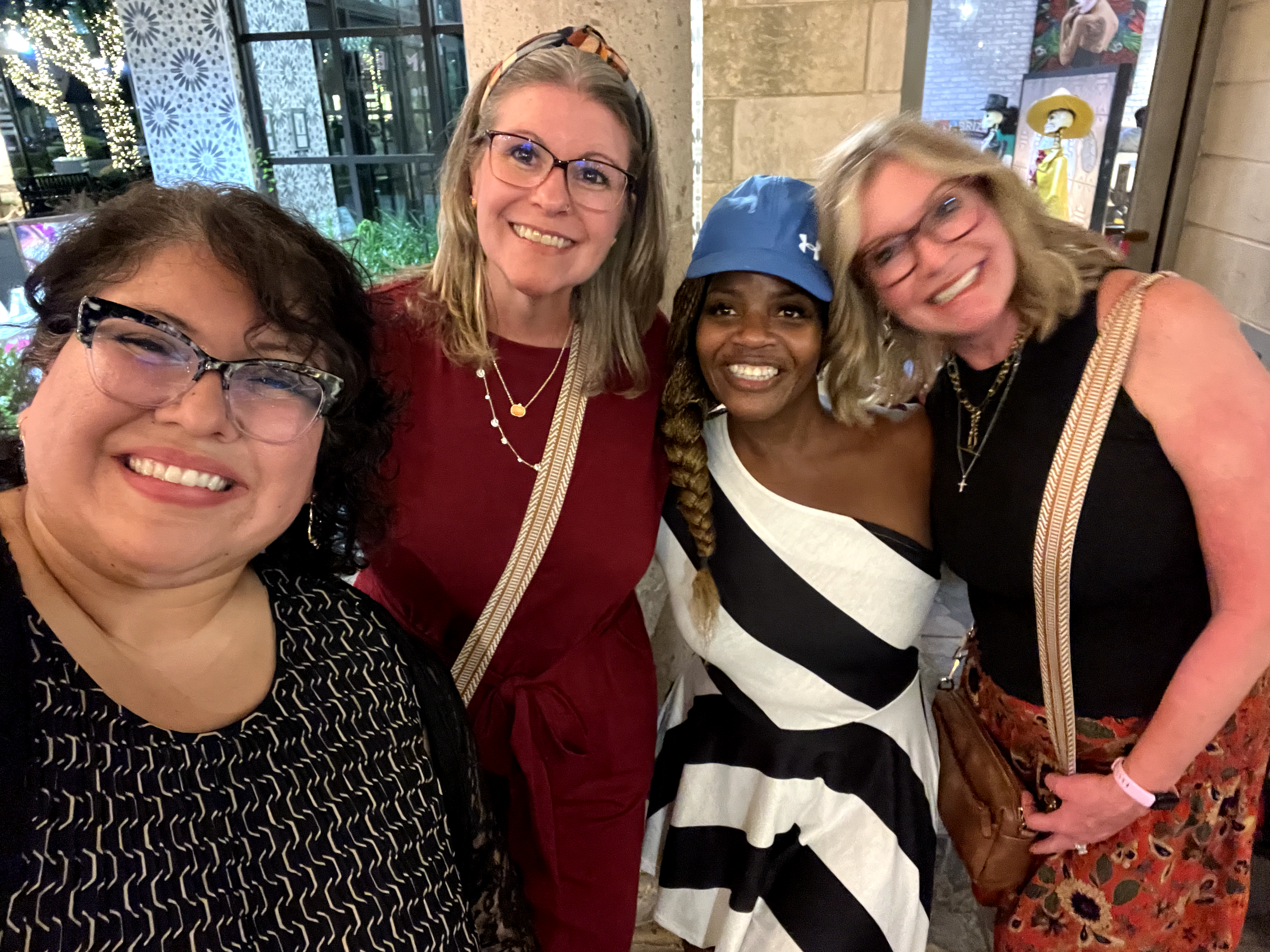 Four women smiling and posing for a photo in an indoor setting with decorative lighting and artwork in the background.