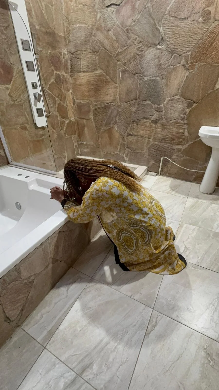 A woman kneeling by a bathtub in a bathroom with large beige and brown stone tiles, wearing a yellow and white patterned dress.