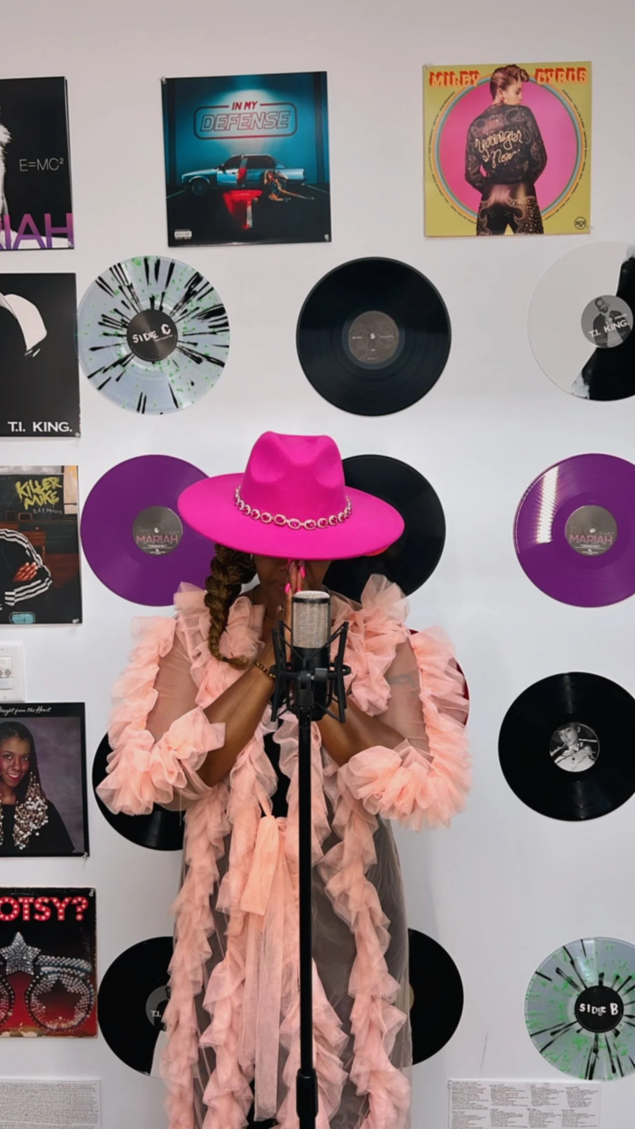 A woman wearing a large pink hat with a chain detail, pink ruffled dress, and long braid, standing in front of a microphone with her hands near her face, against a wall decorated with vinyl records and album covers.