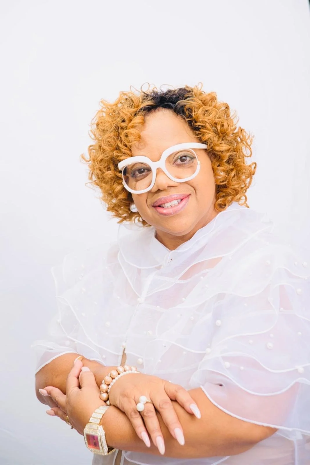 A woman with curly orange hair, wearing large white glasses and dressed in a light, semi-transparent white top with pearl accents, posing with her arms crossed against a plain white background.