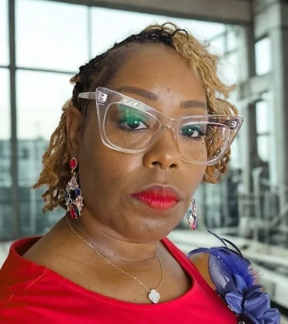 Close-up of a woman with red lipstick, wearing large clear glasses, earrings, and a red top, in a modern indoor setting with glass windows.