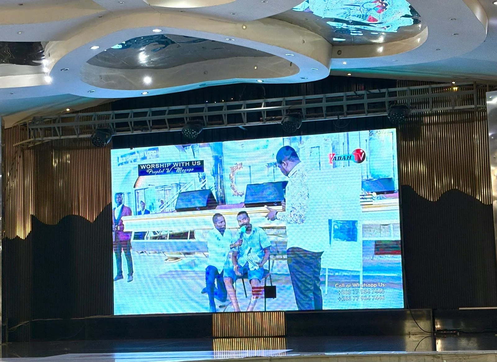 A large digital screen in a venue displaying a church service or event. The screen shows two men sitting, with one man holding a microphone, and a man standing and reading from a tablet. The background of the screen includes a church interior with a