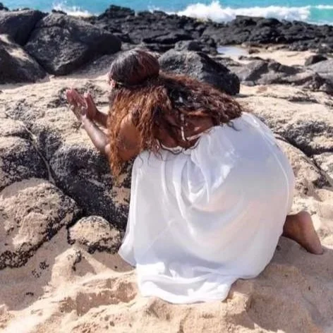A woman in a white dress kneeling on a sandy beach near rocks, with ocean waves in the background.
