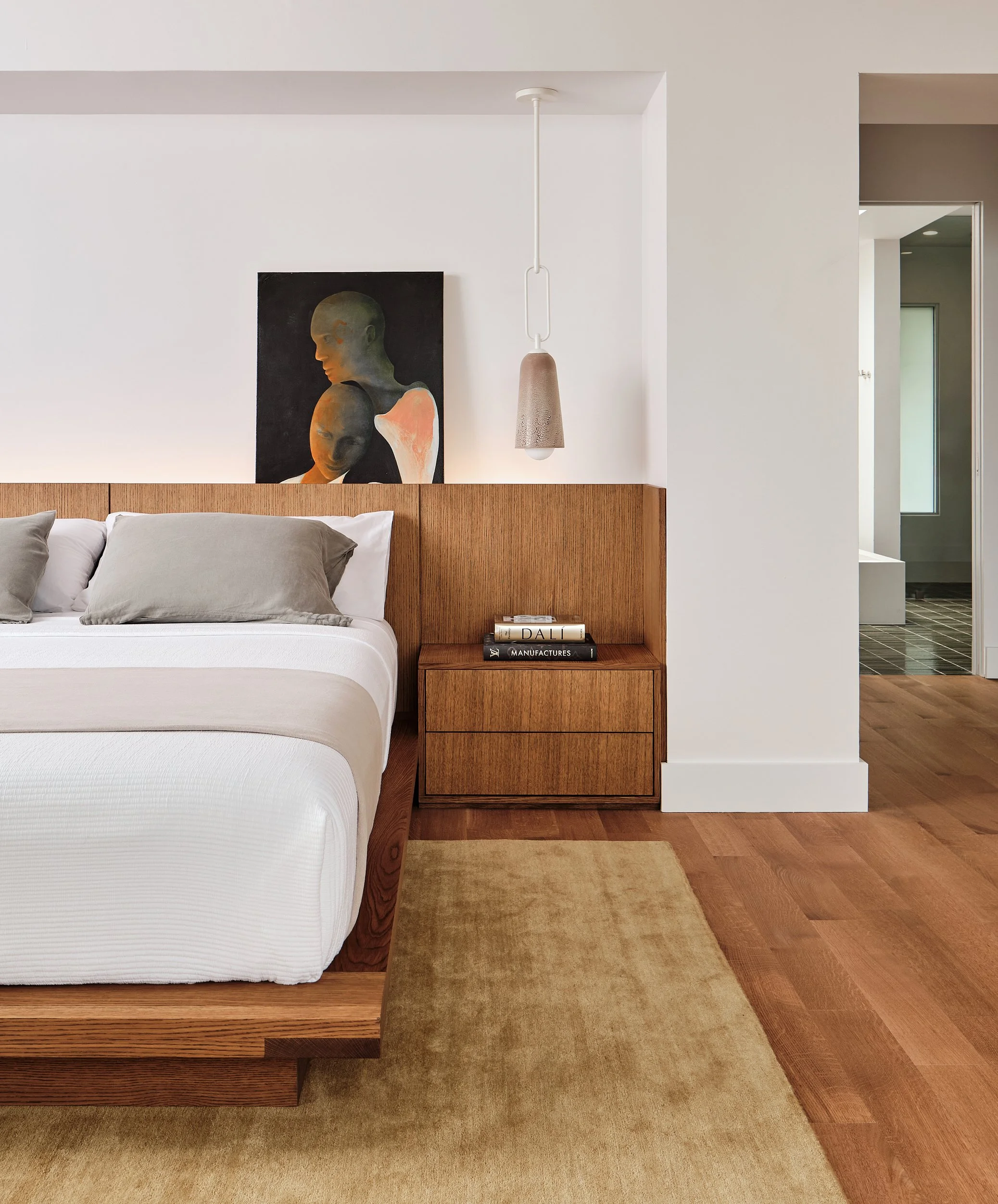 Modern bedroom with a wooden headboard, gray pillows, an abstract portrait painting above the bed, a small wooden nightstand with books, a hanging pendant light, and hardwood floors.