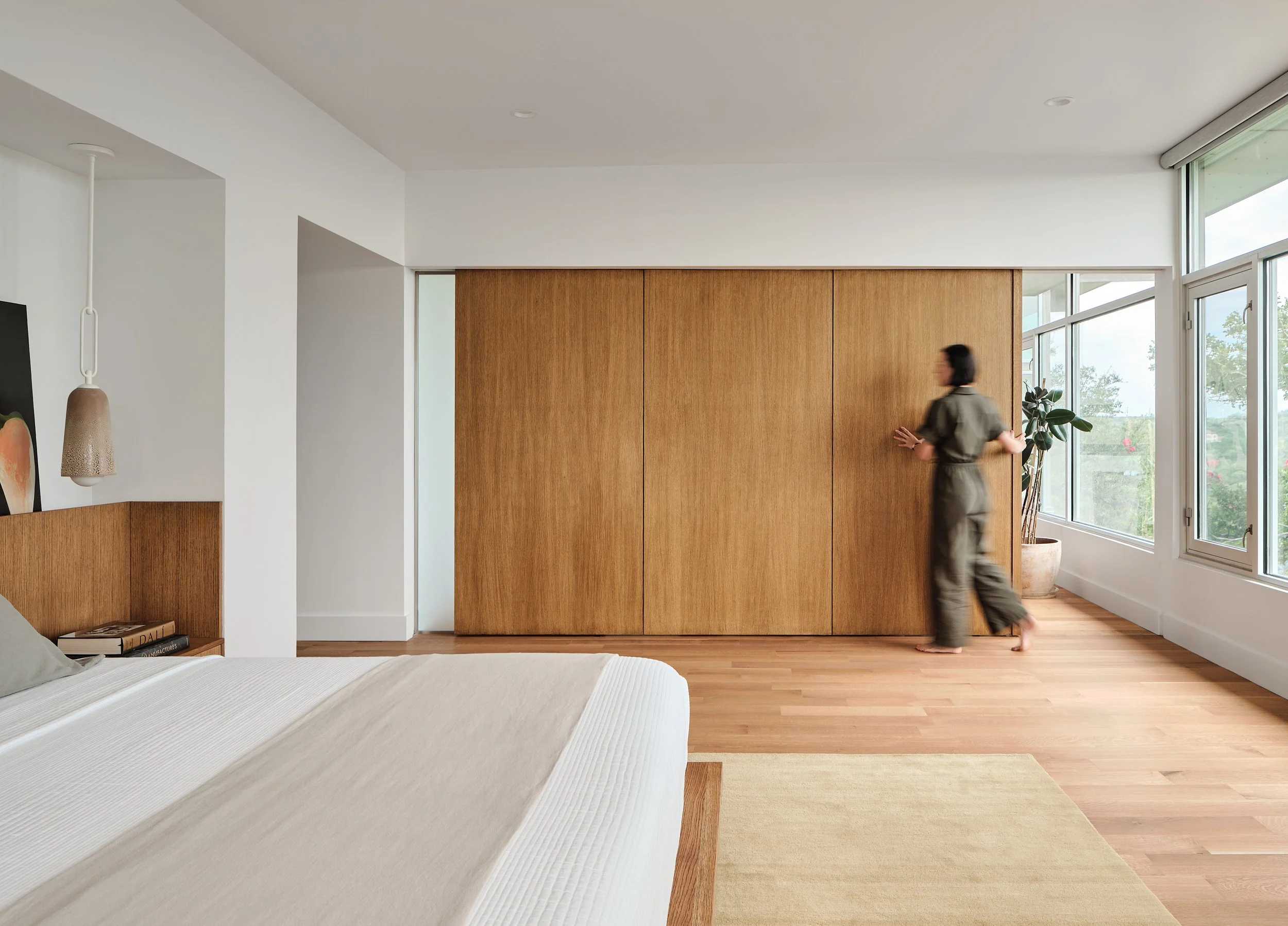A woman with short dark hair in a jumpsuit opening a wooden sliding door in a modern bedroom with large windows and natural light.